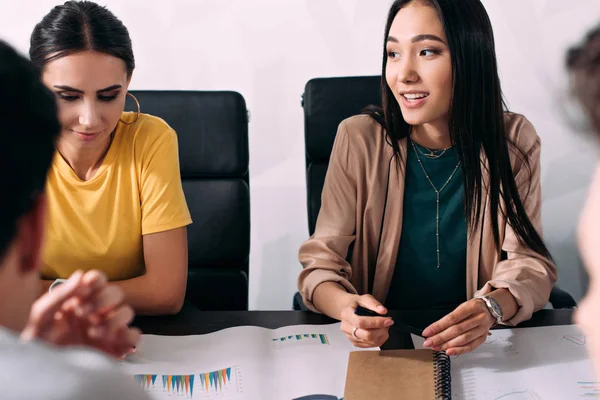 Мультикультурные предпринимательницы сидят за столом с графиками во время деловой встречи в современном офисе — стоковое фото