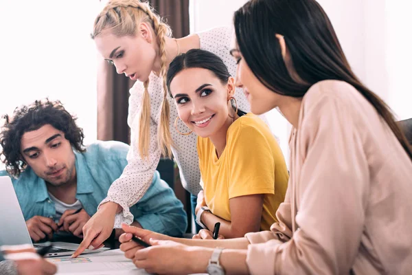 Young businesswoman pointing by finger on graphs to male colleague while two businesswomen talking to each other at modern office — Stock Photo