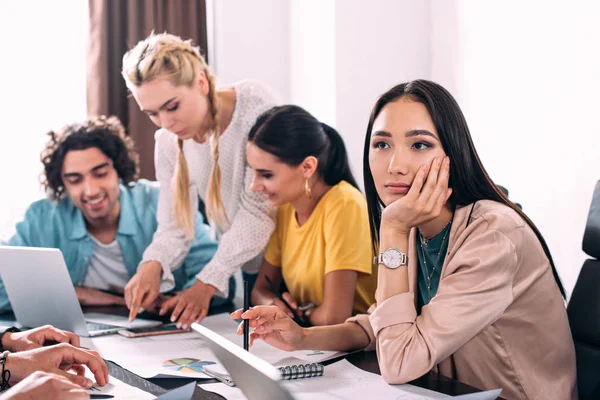 Thoughtful asian businesswoman sitting at table while her colleagues having discussion behind at modern office — Stock Photo