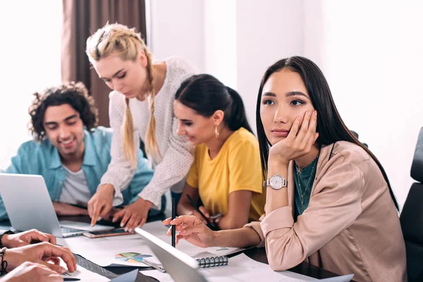 Nachdenkliche asiatische Geschäftsfrau sitzt am Tisch, während ihre Kollegen im modernen Büro hinter der Theke diskutieren — Stockfoto