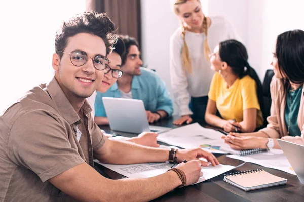 Zwei multikulturelle Geschäftsleute mit Brille blicken in die Kamera, während ihre Partner im modernen Büro im Hintergrund diskutieren — Stockfoto