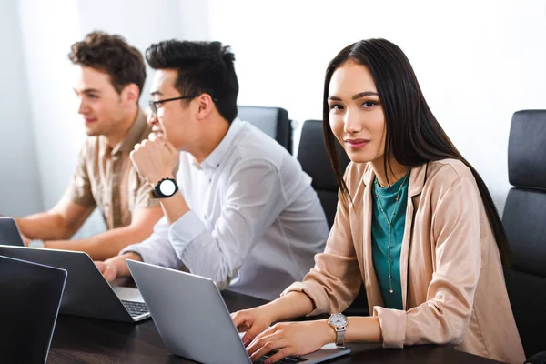Giovane donna d'affari asiatica digitando sul computer portatile mentre i suoi colleghi di avere discussione in ufficio moderno — Foto stock