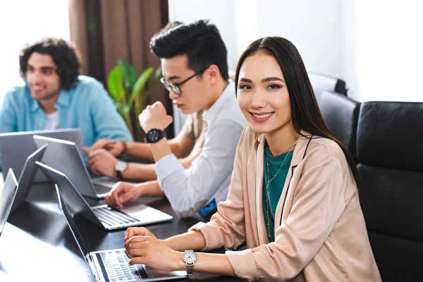 Young asian businesswoman looking at camera at table with laptops and businessmen working behind at modern office — Stock Photo