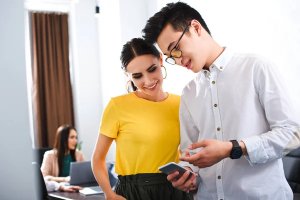 Giovane uomo d'affari asiatico che punta sullo schermo dello smartphone alla donna d'affari sorridente all'ufficio moderno — Foto stock