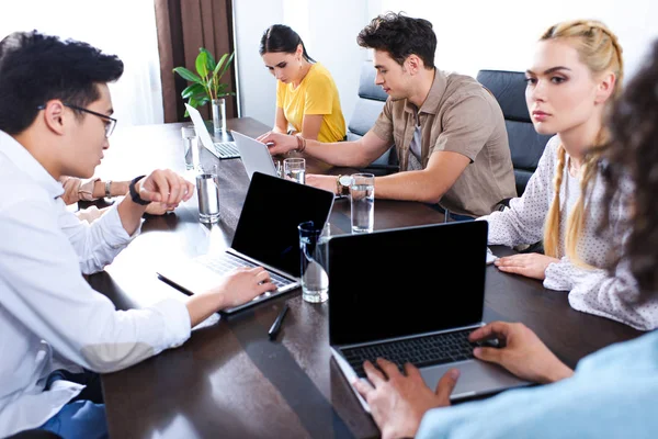 Socios comerciales multiculturales que se reúnen en la mesa con ordenadores portátiles y vasos de agua en la oficina moderna - foto de stock