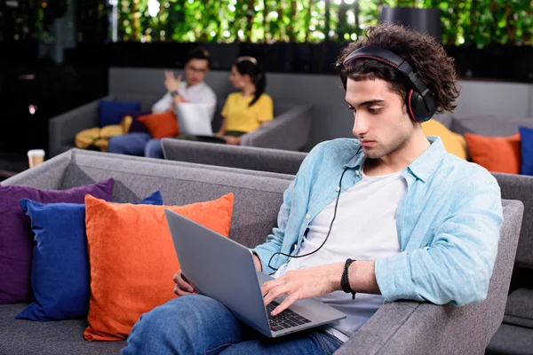 Молодий бізнесмен в навушниках, сидячи на дивані з ноутбуком в сучасному коворкінгу — стокове фото
