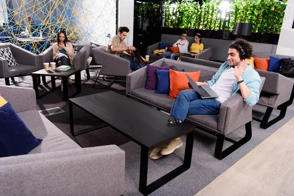 Young businessman in headphones sitting on couch with laptop and waving by hand at modern coworking office — Stock Photo