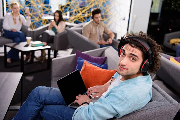 Young businessman in headphones sitting on couch and using laptop with blank screen at modern coworking office — Stock Photo