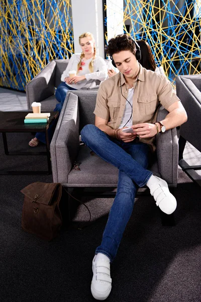Молодий бізнесмен в навушниках зі смартфоном, сидячи на кріслі і бізнес-леді, сидячи позаду з кавою в сучасному коворкінгу — стокове фото