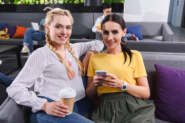 Two smiling businesswomen with coffee and smartphone at modern coworking office — Stock Photo