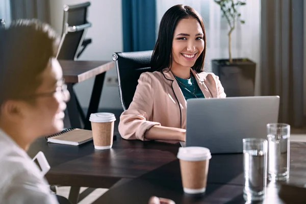 Junge asiatische Geschäftsfrau sitzt mit Laptop, Kaffee und Wassergläsern am Tisch, während ihr Partner im modernen Büro diskutiert — Stockfoto