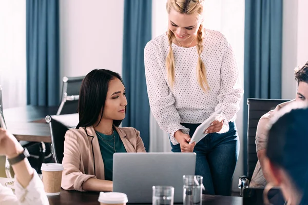 Femme d'affaires souriante montrant tablette numérique à une collègue asiatique tandis que d'autres partenaires ayant des discussions au bureau moderne — Photo de stock