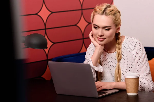 Молода бізнес-леді з паперовою чашкою кави, що сидить за столом з ноутбуком в сучасному офісі — стокове фото
