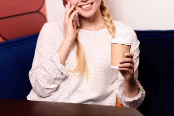 Recortado disparo de sonriente mujer de negocios hablando en el teléfono inteligente y beber café en la oficina - foto de stock