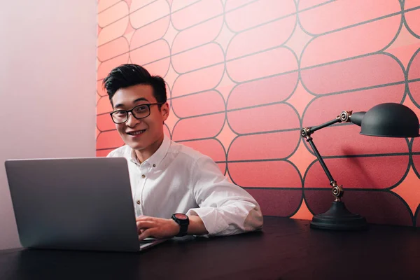Sonriente joven asiático hombre de negocios trabajando en mesa con portátil en oficina - foto de stock
