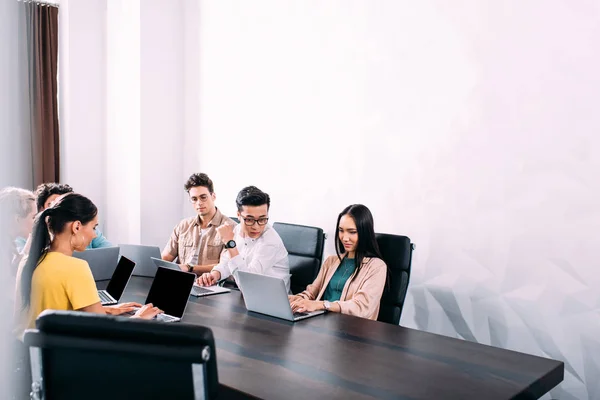 Socios comerciales multiculturales que se reúnen en la mesa con ordenadores portátiles en la oficina moderna - foto de stock