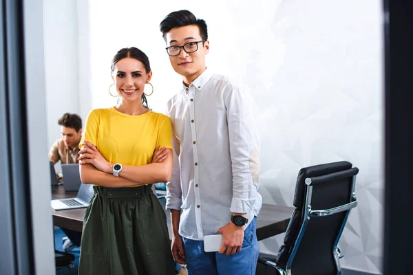 Femme d'affaires souriante et homme d'affaires asiatique avec smartphone regardant la caméra au bureau moderne — Photo de stock