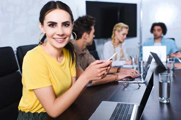 Giovane donna d'affari sorridente con smartphone e i suoi partner che discutono dietro al moderno ufficio — Foto stock