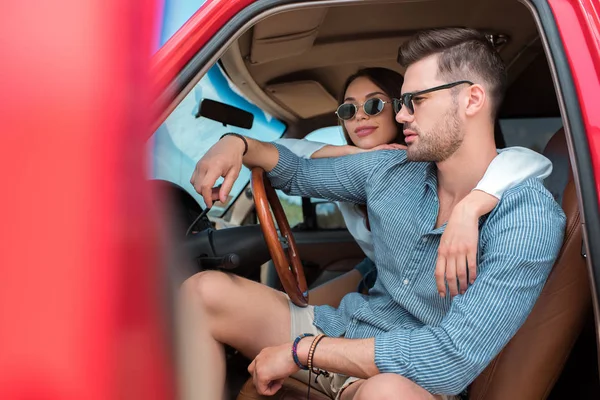 Jovem casal de viajantes em óculos de sol sentado no carro e abraçando juntos — Fotografia de Stock