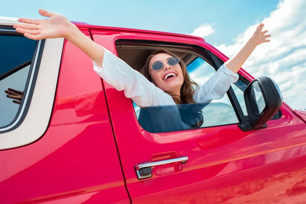 Chica excitada en gafas de sol haciendo gestos y sentado en coche rojo durante el viaje por carretera - foto de stock