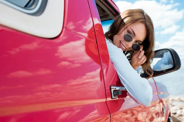 Hermoso viajero sonriente en gafas de sol sentado en coche rojo durante el viaje por carretera - foto de stock