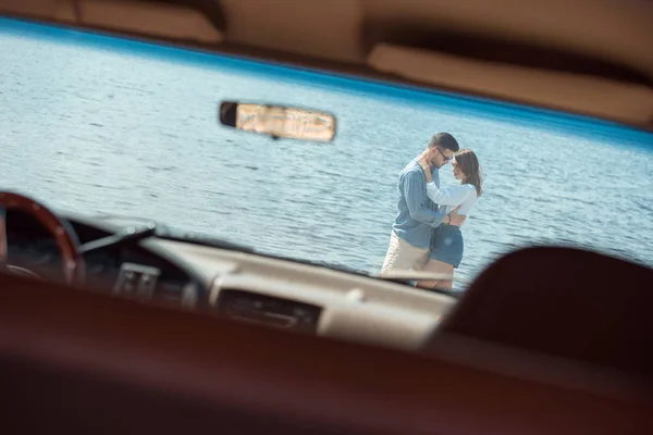 Молодая пара путешественников обнимаются у моря, вид через автомобиль — стоковое фото