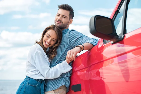 Веселая молодая пара, обнимающаяся возле красной машины — стоковое фото