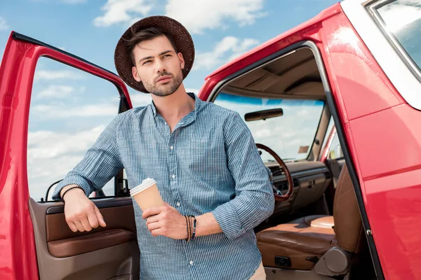 Bel homme au chapeau tenant du café pour aller et debout à la voiture pendant le voyage sur la route — Photo de stock