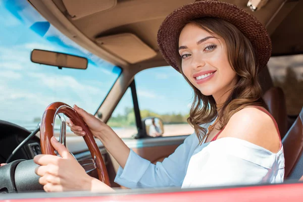 Привлекательная улыбающаяся женщина в шляпе за рулем автомобиля во время дорожной поездки — стоковое фото