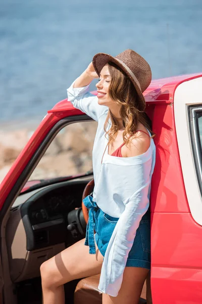 Attraktive Frau mit Hut posiert bei Roadtrip in der Nähe des Meeres vor rotem Auto — Stockfoto