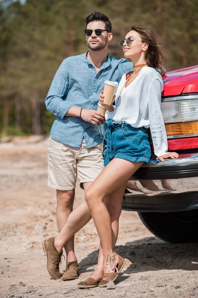 Casal de viajantes com copos descartáveis de café de pé no carro — Fotografia de Stock