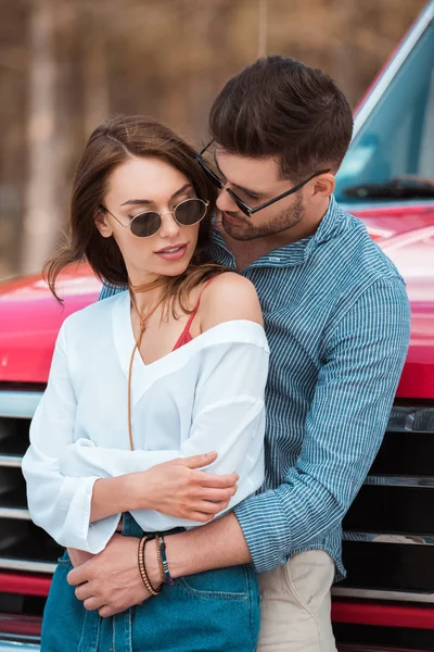Tendre beau couple en lunettes de soleil étreignant près de voiture rouge — Photo de stock