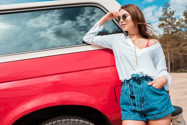 Chica atractiva en gafas de sol posando cerca de coche rojo durante el viaje por carretera — Stock Photo