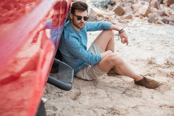 Bello uomo elegante in occhiali da sole seduto vicino auto rossa — Foto stock