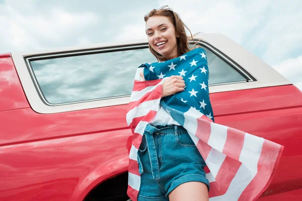 Cheerful girl wrapped in american flag standing near red car — Stock Photo