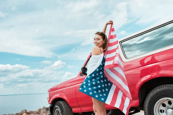 Menina bonita feliz com bandeira americana de pé perto do carro vermelho — Fotografia de Stock