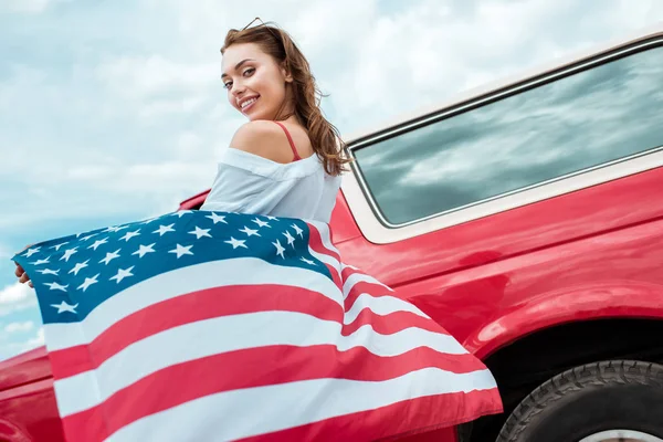 Menina atraente com bandeira americana de pé perto de carro vermelho — Fotografia de Stock