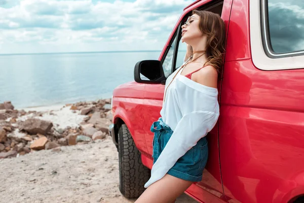 Alegre joven mujer posando ar ar coche rojo durante el viaje cerca del mar - foto de stock