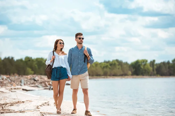 Happy couple holding hands and walking with backpacks on sea shore — Stock Photo