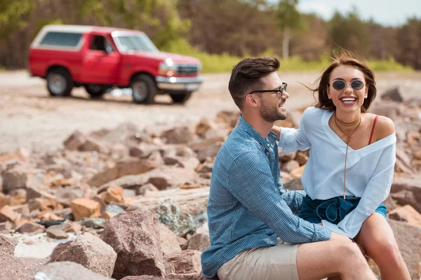 Sonriente pareja sentado en rocas con rojo jeep en el fondo - foto de stock