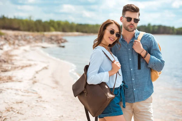Hermosa pareja feliz en gafas de sol con mochilas abrazando en la orilla - foto de stock