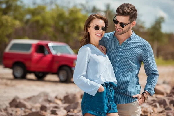 Giovane coppia sorridente in occhiali da sole che si abbracciano insieme, auto sullo sfondo — Foto stock