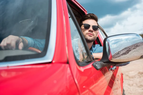 Hombre guapo en gafas de sol sentado en coche rojo durante el viaje por carretera - foto de stock
