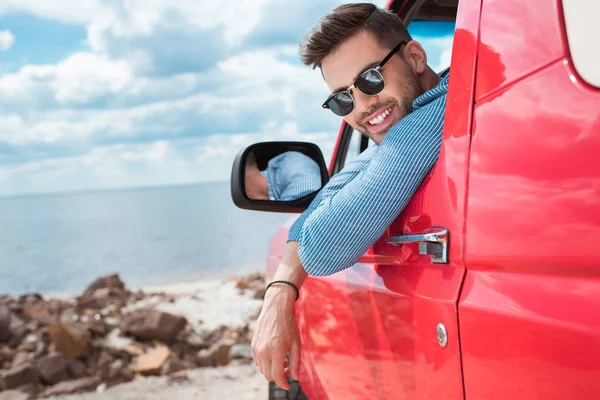Bonito alegre homem em óculos de sol sentado em carro vermelho durante viagem de carro perto do mar — Fotografia de Stock