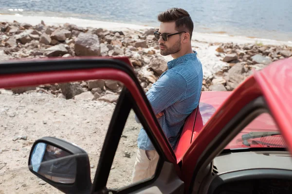 Красивий стильний мандрівник, що стоїть на машині біля моря під час подорожі — стокове фото