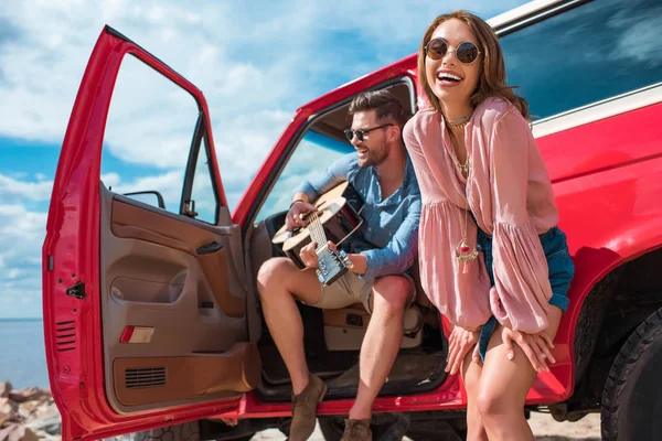 Joven tocando la guitarra cerca de coche con alegre novia - foto de stock