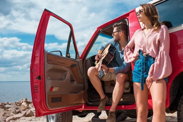 Young man playing acoustic guitar near car with girlfriend on road trip — Stock Photo