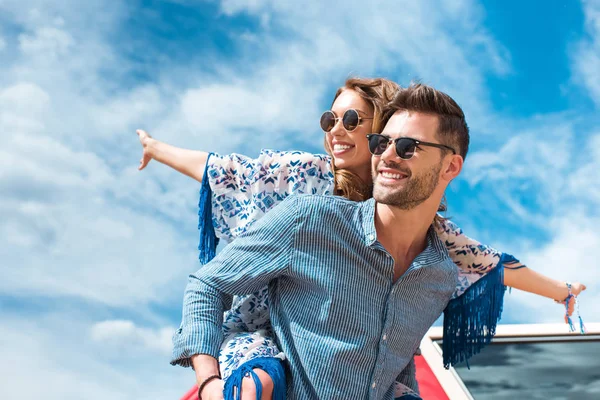 Feliz hombre piggybacking su novia sonriente con el cielo azul en el fondo - foto de stock