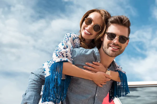 Bel homme en lunettes de soleil piggyback sa petite amie souriante — Photo de stock