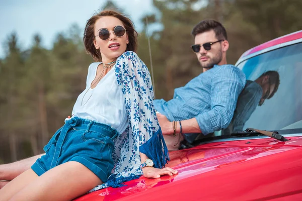 Молода стильна пара в сонцезахисних окулярах сидить на червоній машині — стокове фото